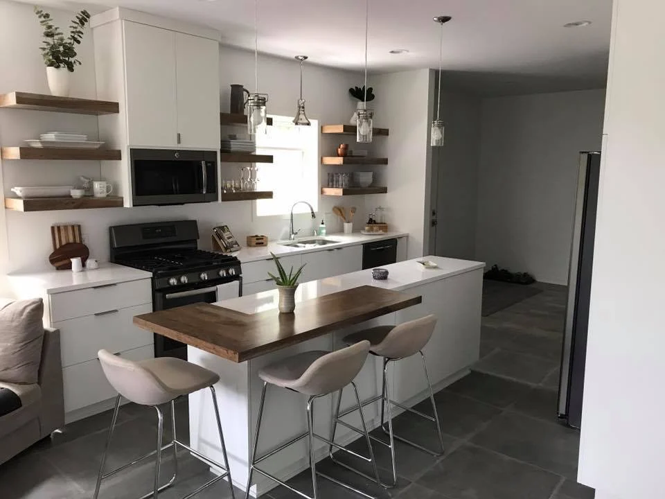 A modern white kitchen with floating wood shelves, a butcher block peninsula, bar stools, and pendant lights.
